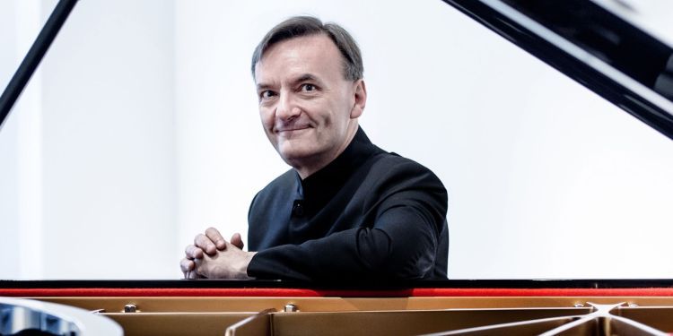 A smiling Stephen Hough wearing a black suit sat behind a piano with a white backdrop