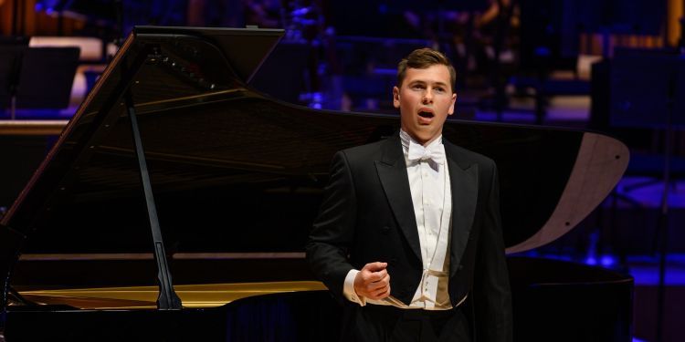 Male presenting student wearing a suit on stage singing with a piano in the background