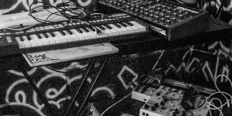 Close-up of an electronic music setup, including a small MIDI keyboard, a synthesizer with multiple knobs labeled "Sequential," and various audio cables and effects units against a graffiti-style background.