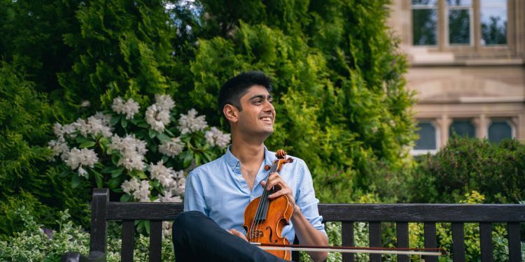 Ammal Bhatia sitting on a bench outdoors, holding a violin and smiling.