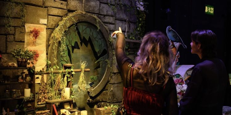 People are stood with their back to the camera looking and pointing to a piece of set with a smashed circular window in the middle and candles and herbs decorating the edge.