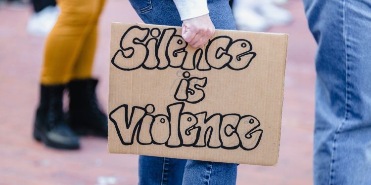 Someone holding a cardboard sign that spells out 'Silence is violence' by their legs