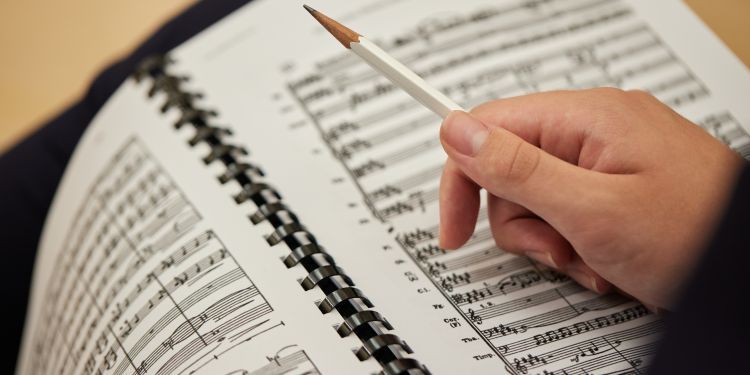 Close-up of a spiral-bound music score book with a hand holding a pencil above the pages, appearing to make notes or analyse the orchestral sheet music.