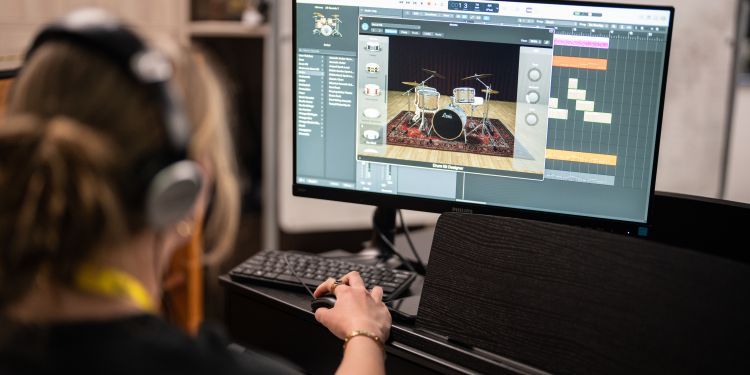 Person wearing headphones sitting at a desk, using music production software on a computer. The screen shows a virtual drum kit and a sequencer with multiple tracks.