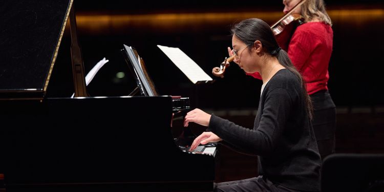 A student playing the piano with a student playing the violin in the background