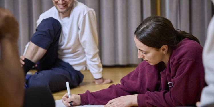 Person lying on the floor writing on a large sheet of paper, with another person seated nearby in a casual indoor setting.