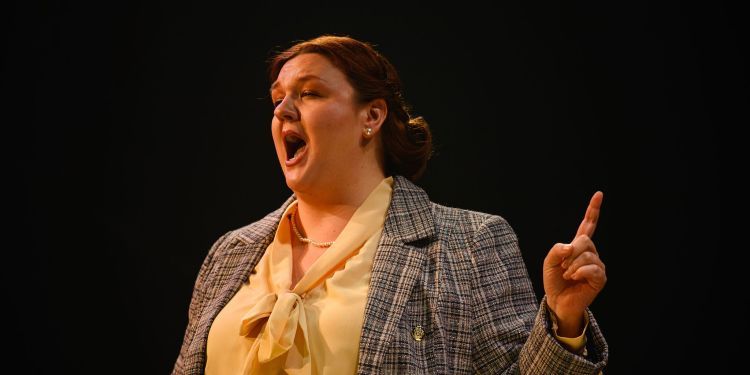 Female presenting singer on stage wearing a checked blazer and white blouse holding their finger up pointing