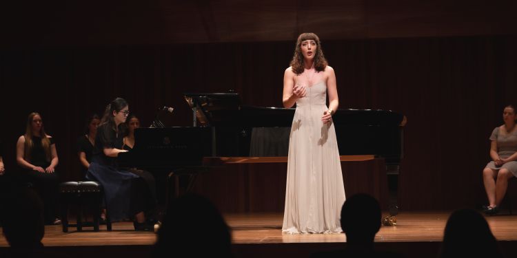 Person in a white gown performing on stage beside a grand piano, with audience silhouettes in foreground.