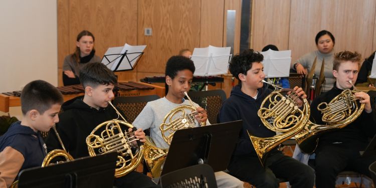 Five young male horn players, sat down, playing their instruments