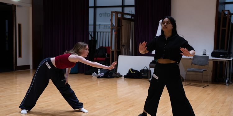 Two people are engaged in a movement exercise in a spacious studio with wooden floors, one leaning forward with an outstretched arm while the other stands with knees bent and hands raised in a grounded stance.