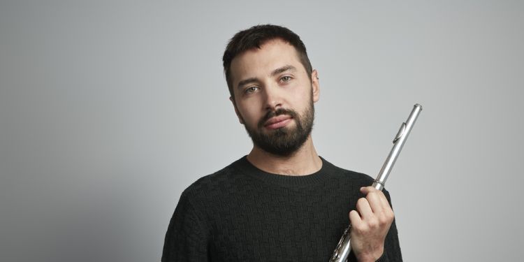 Adam Walker holding a flute looking at the camera in front of a grey background