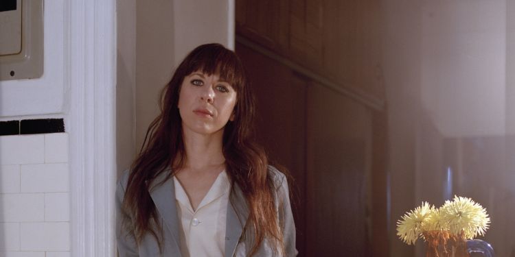Missy Mazzoli, a woman with red hair leans against a door wearing a grey suit