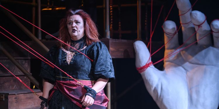 A woman sings whilst connected to red strings and a giant hand