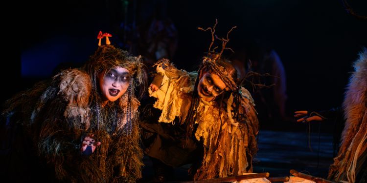 Two woodland creatures, covered in moss and sticks are huddled around four lanterns on sticks which are lit up, causing an orange glow on their faces.