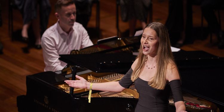 Female presenting singer on stage wearing a floor length black evening gown singing in front of a piano