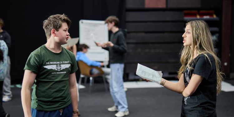 two drama short courses participants acting in a room holding a piece of paper with other participants in the background