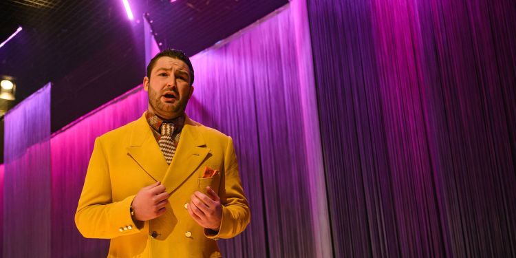 Opera student wearing a yellow suit singing on stage with a pink fringe backdrop