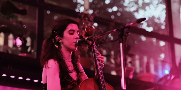 A musician performs live on stage, singing into a microphone while playing the cello under pink lighting.