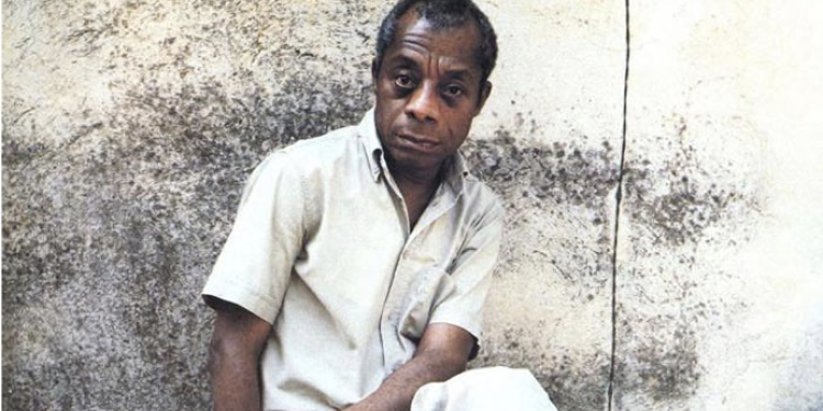 James Baldwin wearing a white shirt smiling at the camera in front of a concrete wall