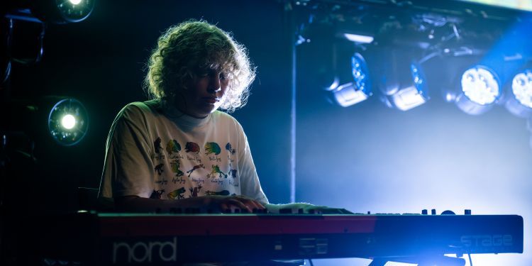 A musician playing a Nord keyboard under blue stage lights, with a cosmic backdrop