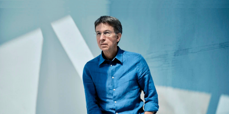 Man sitting on a stool, dressed in blue top and beige pants. 