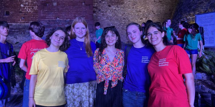 Five young women smiling together outdoors at night in front of an illuminated historic stone wall, with several wearing matching London Schools Symphony Orchestra t-shirts.
