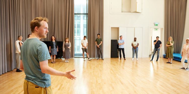 A man in the foreground with his arms reached out talks to a room of people in a circle in a training room