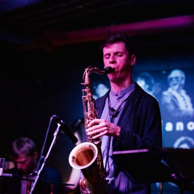A man wearing a dark blazer and t-shirt plays a saxophone on-stage. There are projections in the background
