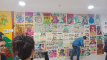 wall displaying many colourful posters with a person pasting one to the wall and another taking a photograph of the wall