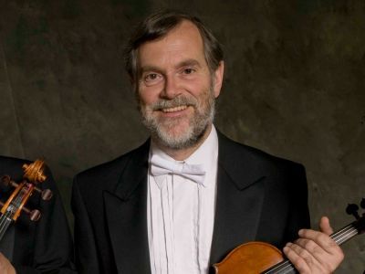 A man stands in his suit holding a violin. He has a smile on his face