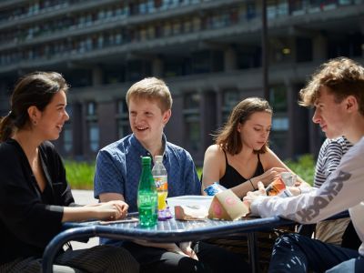 From left to right: A girl with a black shirt, boy with a blue shirt, girl with black top and boy with white top are sat eating lunch at a table.