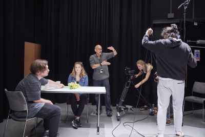 A male and female actor are sat at a table preparing to shoot a scene. The director is stood behind them, using his arms to instruct the camerawoman and boom operator how to frame the shot.