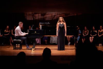 A young woman stands on stage, singing to an audience in a long black dress.