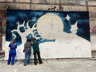 Students painting a stage backdrop with ghostly moon and trees