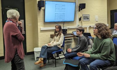 Three young students sit in a classroom and to the left Dame Judith Weir stands talking to them