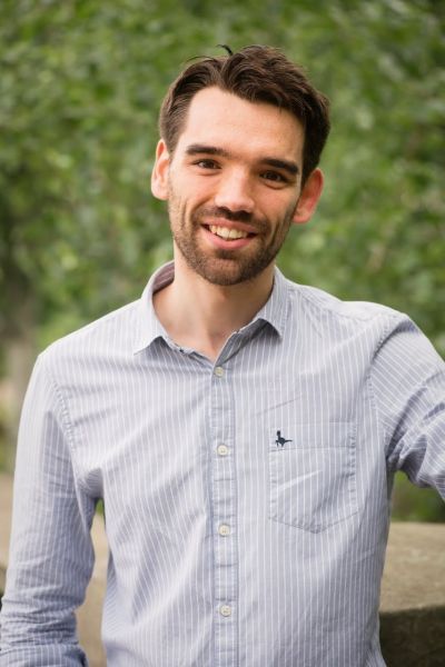 A head and shoulders portrait of a young man with brown hair and beard smiling at the camera. He is in an outdoor setting with greenery behind him