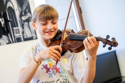 A girl with a blond fringe and hair tied back smiles while playing the violin in a room with white walls and a black and white picture hung up behind her