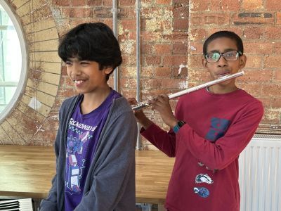 Two young boys, the one on the left is smiling and playing the keyboard, the one on the right is playing the flute, they are in a well lit room with red brick wall