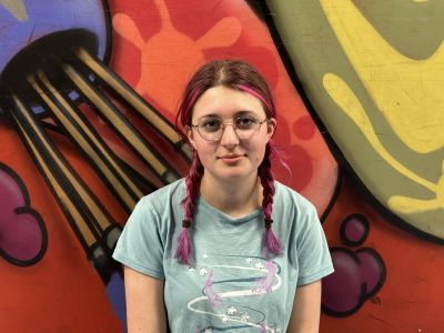 A girl with two red plaits and a green t-shirt smiles at the camera, behind her is a colourful painted wall
