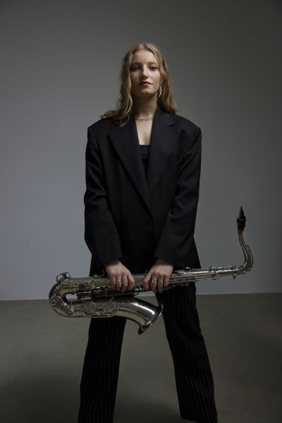 Female blond saxophonist standing in black suit holding instrument