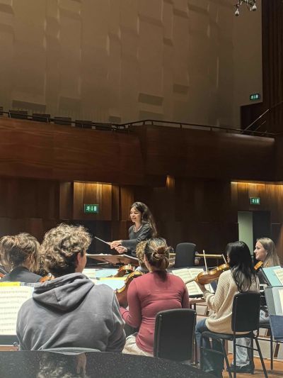 A conductor leads a seated string ensemble during a rehearsal in a wood-panelled concert hall.