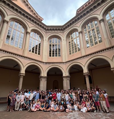Large group of students posing together in an outdoor courtyard surrounded by classic arched columns and large arched windows.