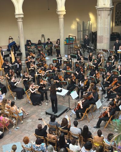 A student orchestra performs in a courtyard, conducted from a central podium and surrounded by a seated audience.