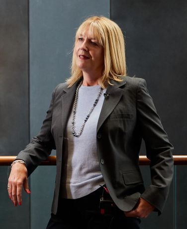 A woman with shoulder length blond hair looks off camera, she is wearing a grey suit